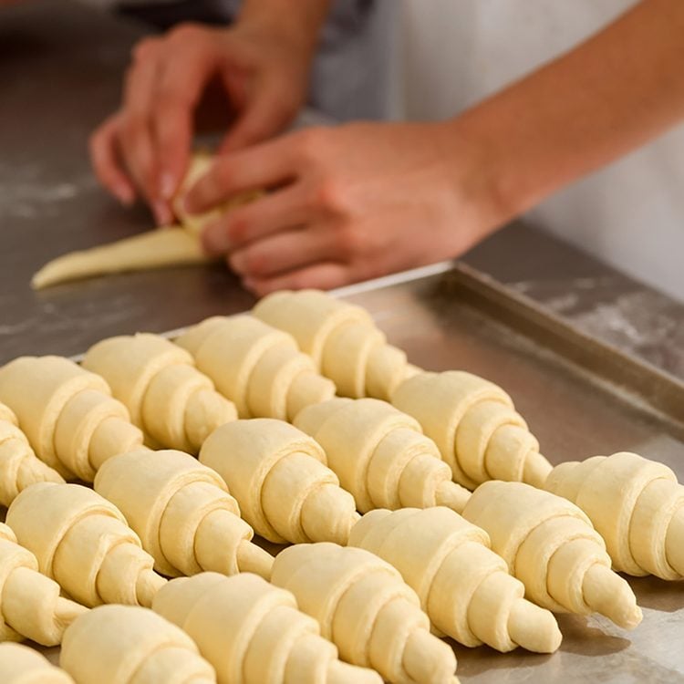 Croissants crus façonnés en rangs sur un plan de travail, et deux mains en train de rouler un triangle de pâte levée feuilletée